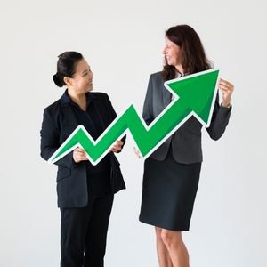 Two businesswomen smiling while holding a large upward arrow sign, symbolizing growth and success in their careers.